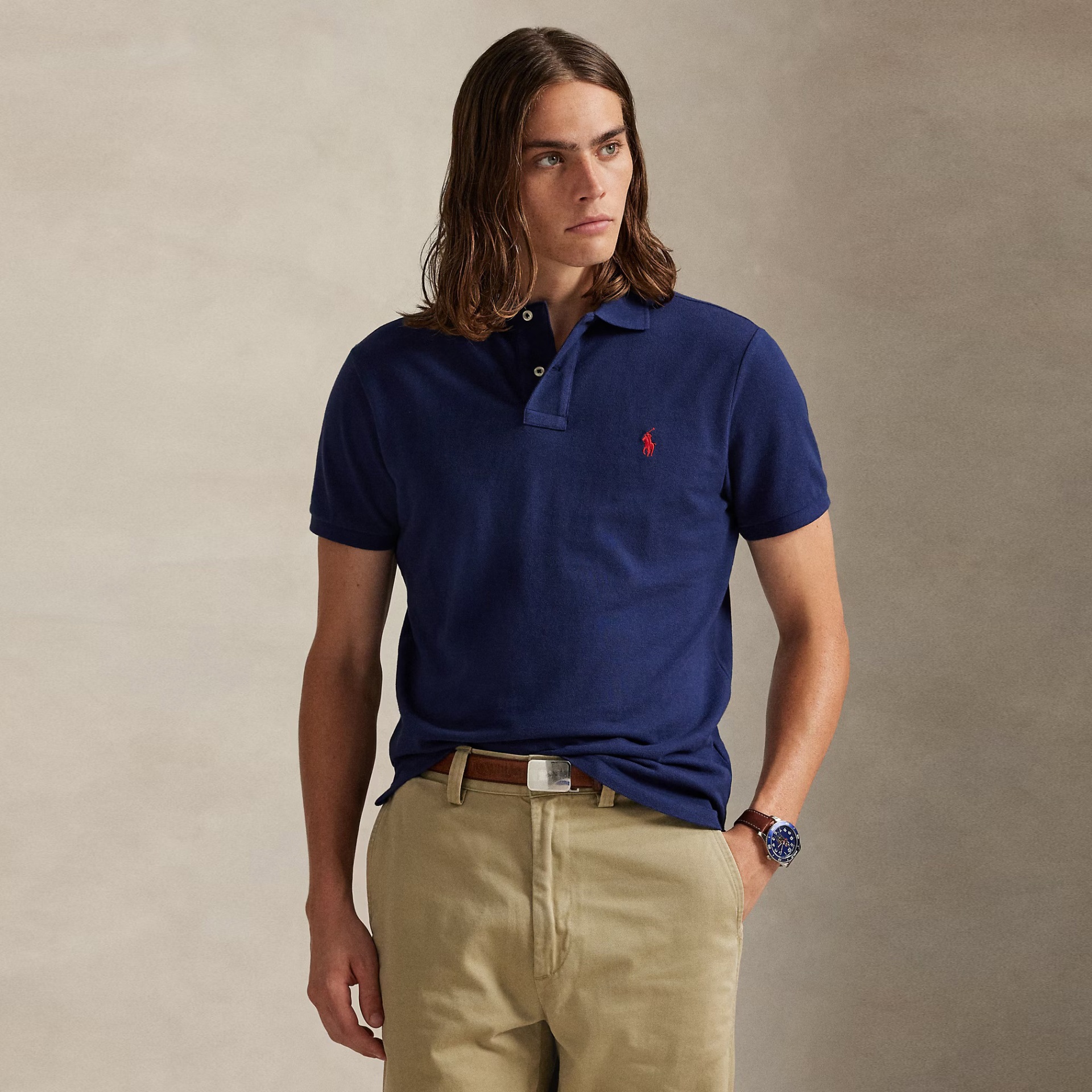 Homem com polo azul escuro, calças cáqui e relógio, fundo neutro.