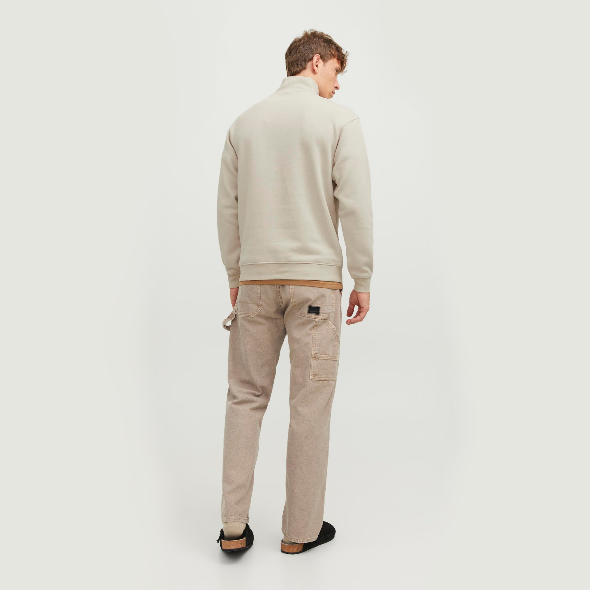 Homem de costas com casaco bege claro e calças bege, calçado preto e castanho, fundo branco