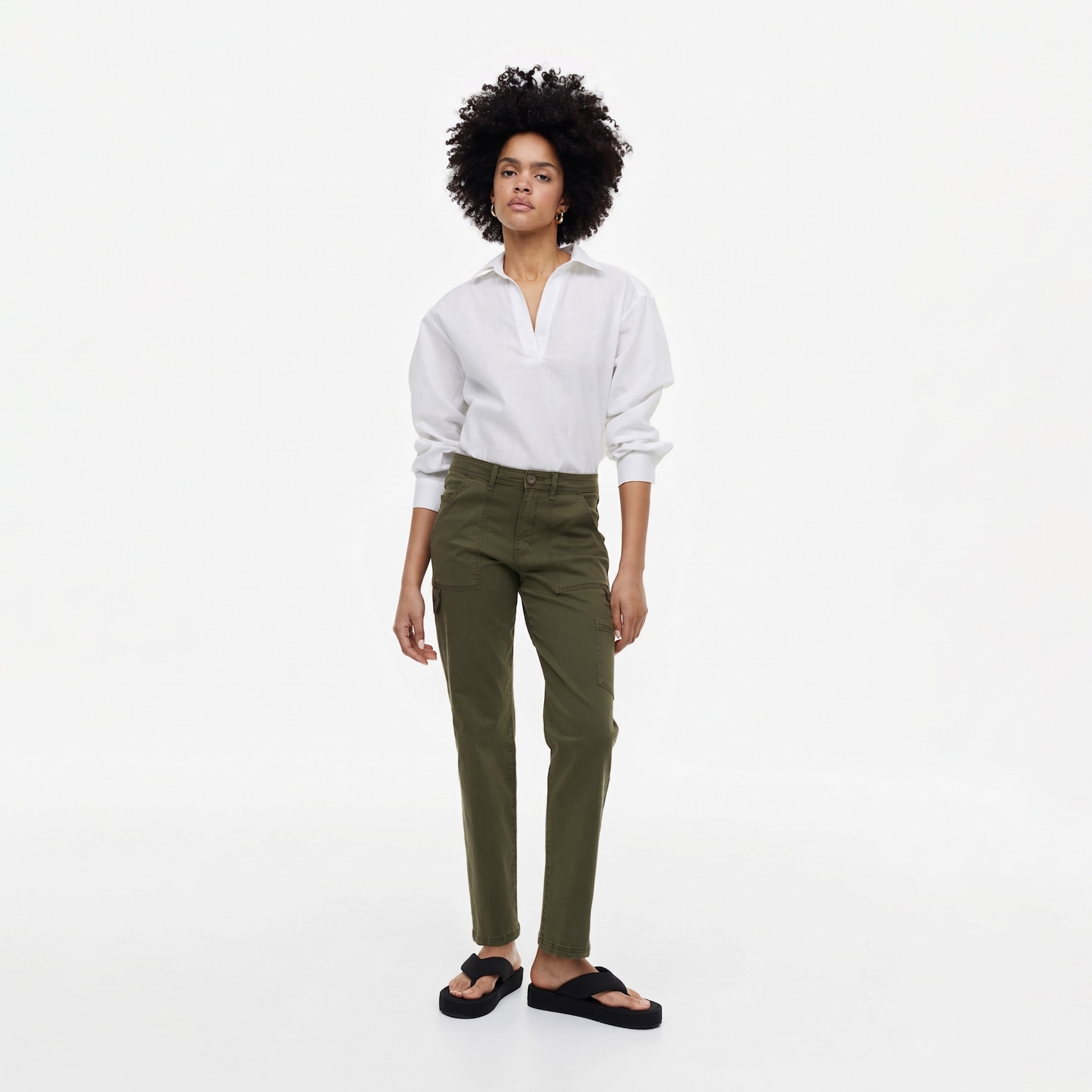 Mulher vestindo pantalonas cargo verdes, sapatos pretos de plataforma e camisa branca em fundo branco