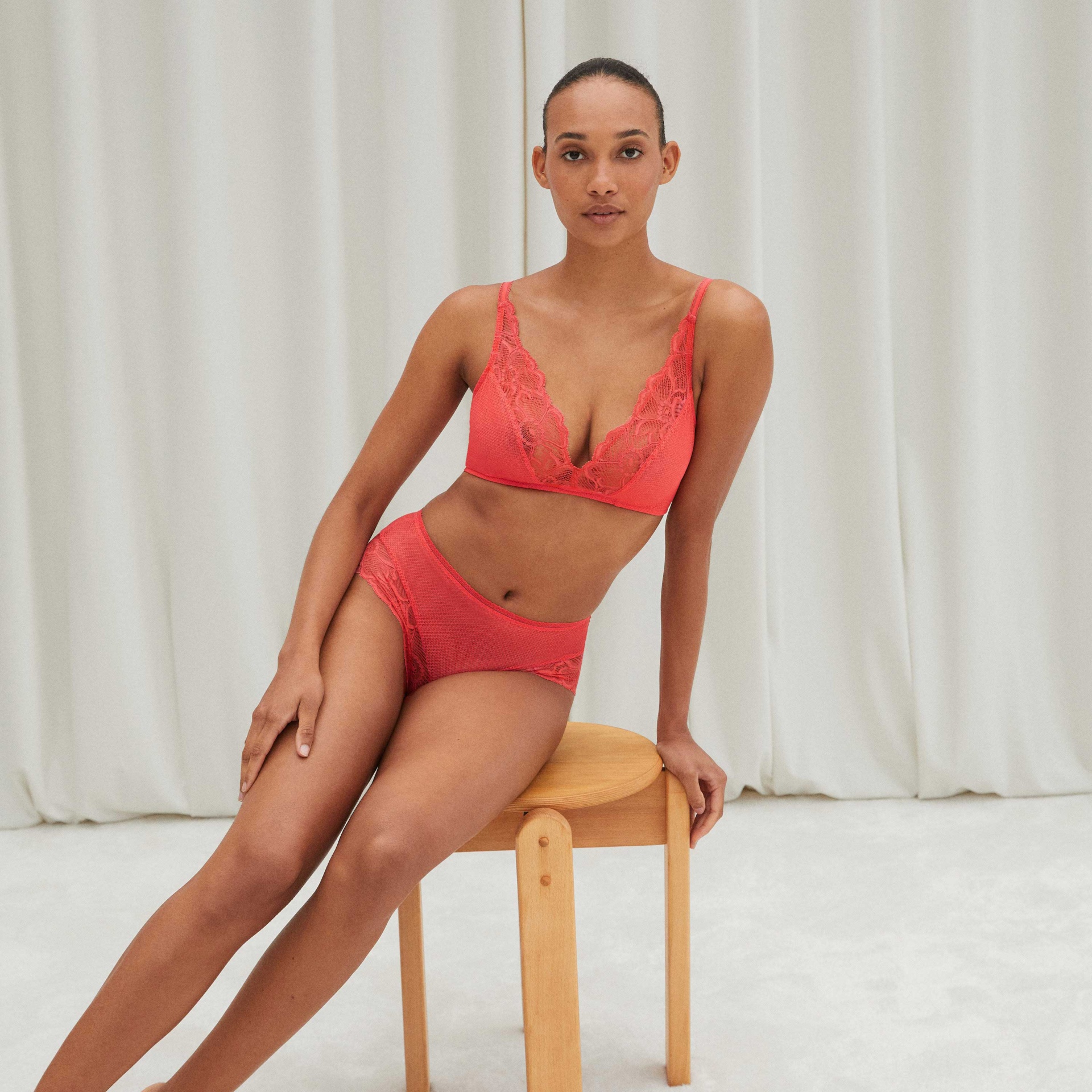 Conjunto de roupa interior vermelho com renda em fundo branco modelo sentada num banco de madeira