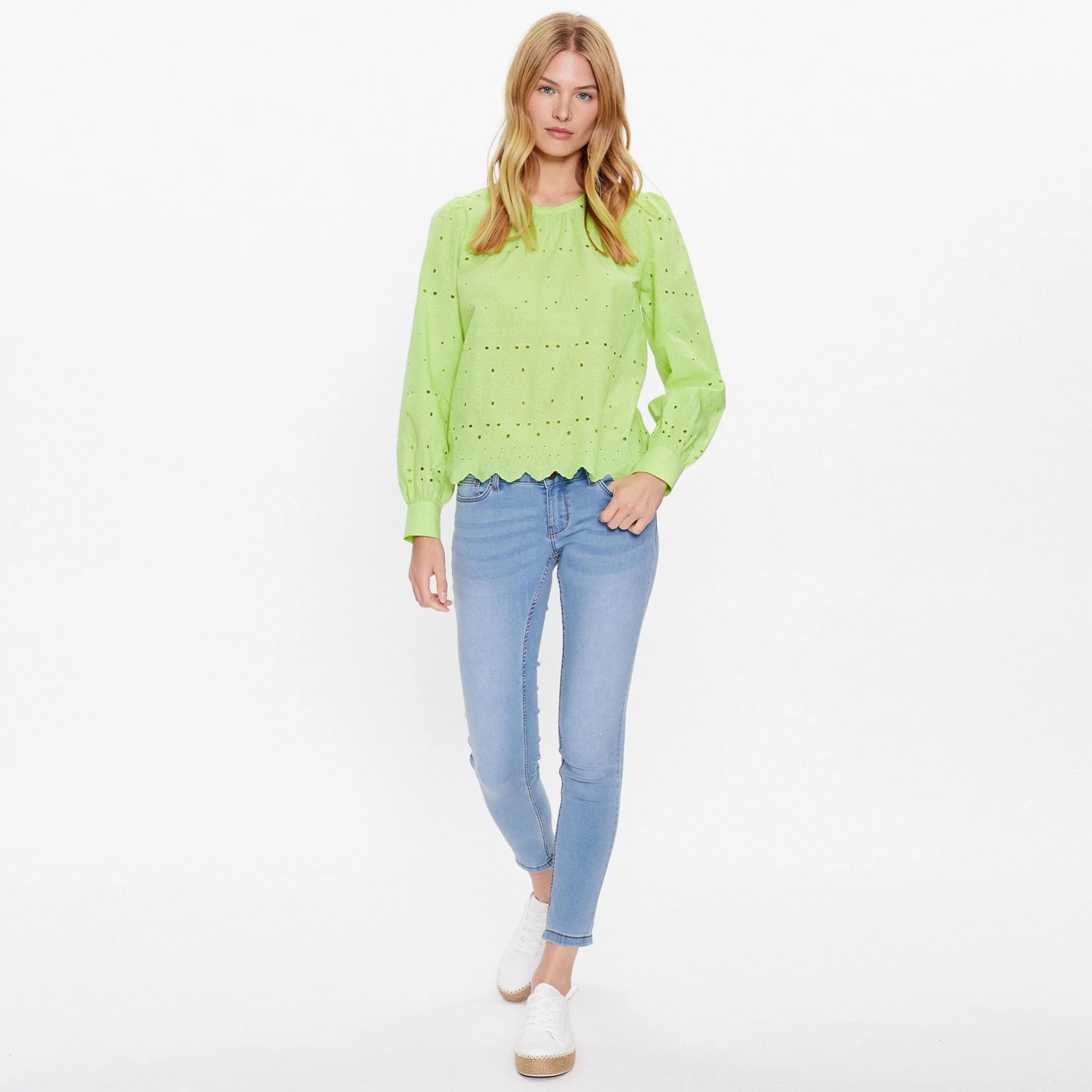 Mulher vestindo camisola verde clara com padrão de pequenos orifícios, calças justas azuis claras e sapatos brancos com fundo branco.