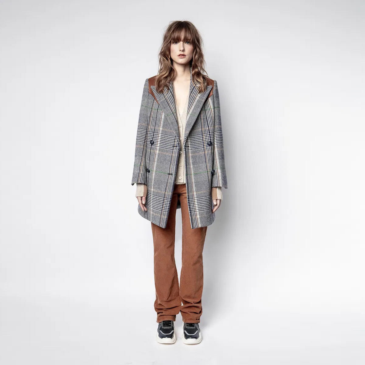Mulher a vestir casaco de padrão aos quadrados com ombros castanhos, camisola bege, calças castanhas e sapatilhas pretas.