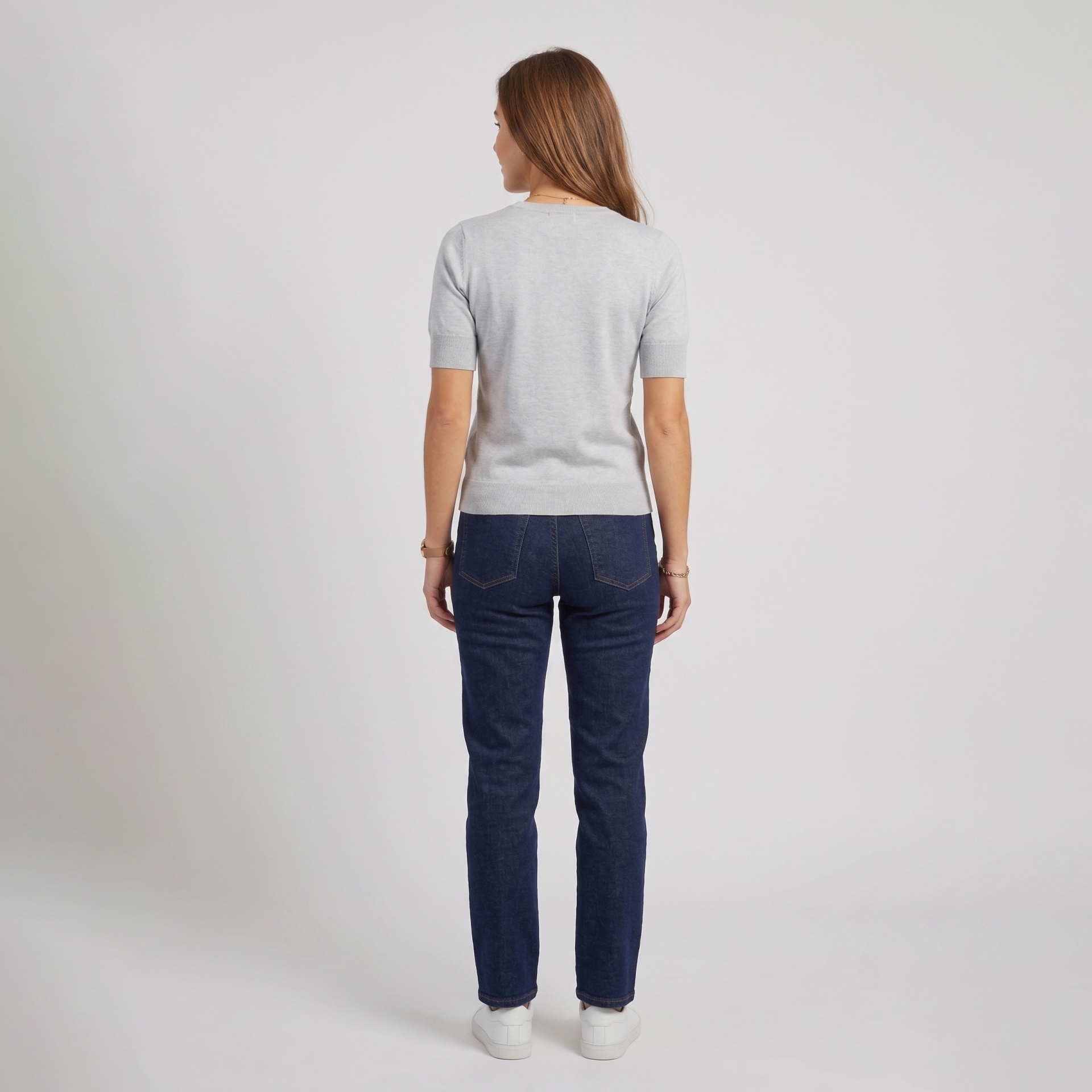 Mulher de costas com camisola cinza, calças de ganga azul escuro e ténis brancos