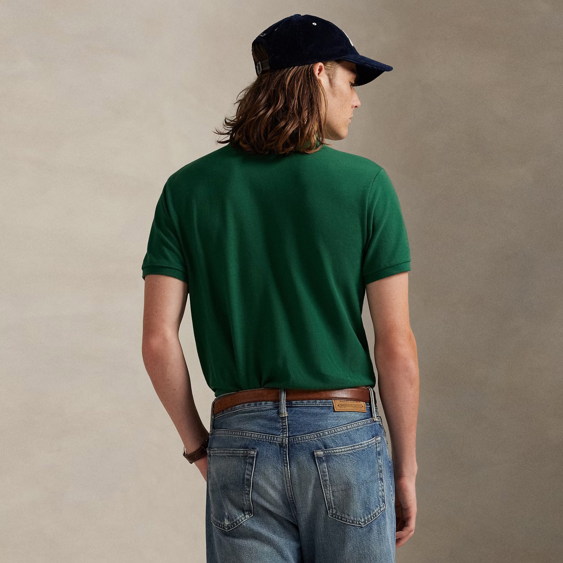 Homem de costas com polo verde, jeans desbotados, cinto castanho e boné preto