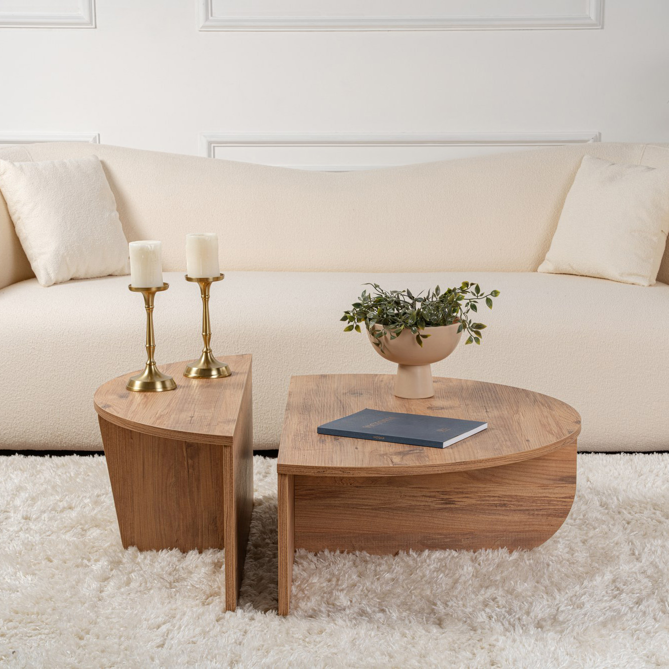 Mesa de centro dupla em madeira clara com castiçais, vaso com planta e livro sobre tapete felpudo, sofá creme ao fundo