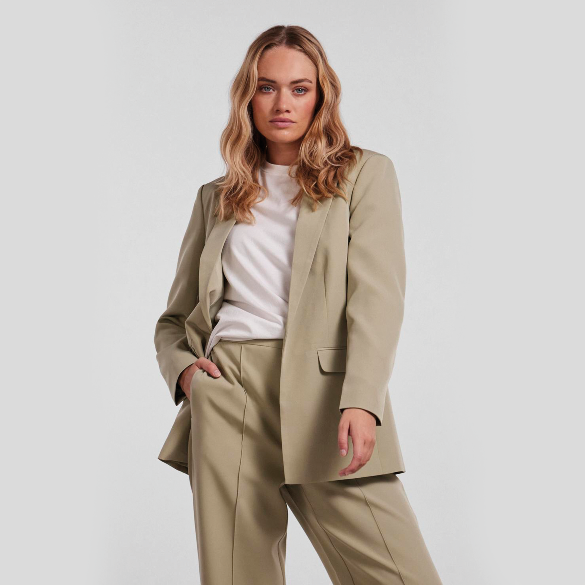 Mulher vestindo terno bege com blazer e calças, t-shirt branca e fundo cinzento