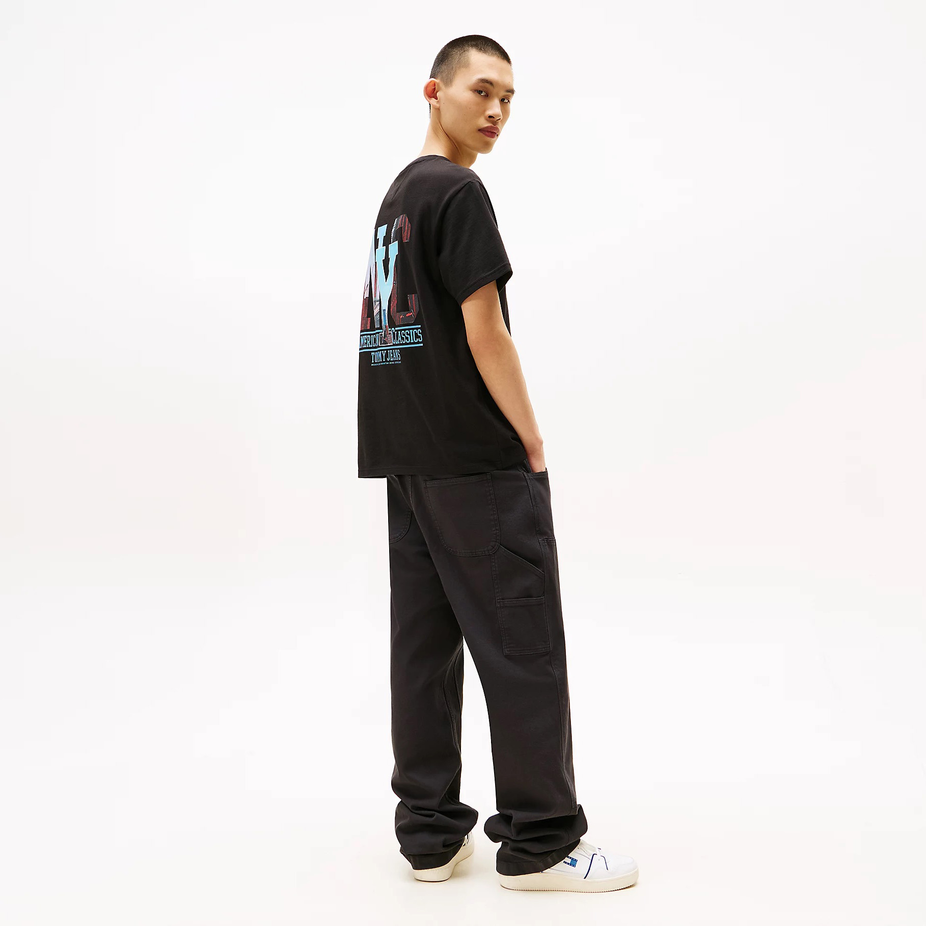 Homem jovem de pé vestido com t-shirt preta estampada, calças pretas largas e ténis brancos, fundo branco.