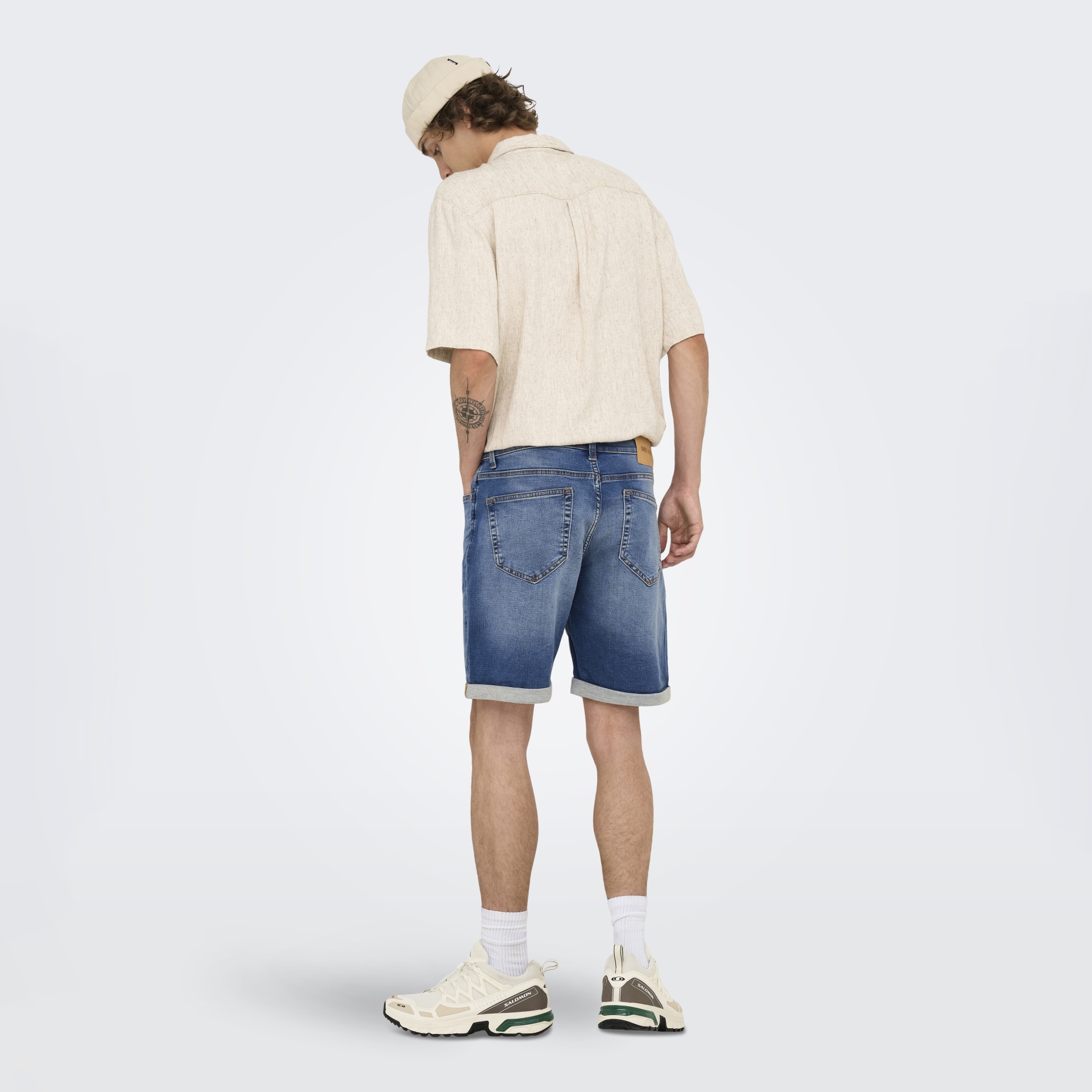 Homem de costas com camisa bege, calções de ganga azuis, meias brancas, sapatilhas brancas Salomon e gorro bege.