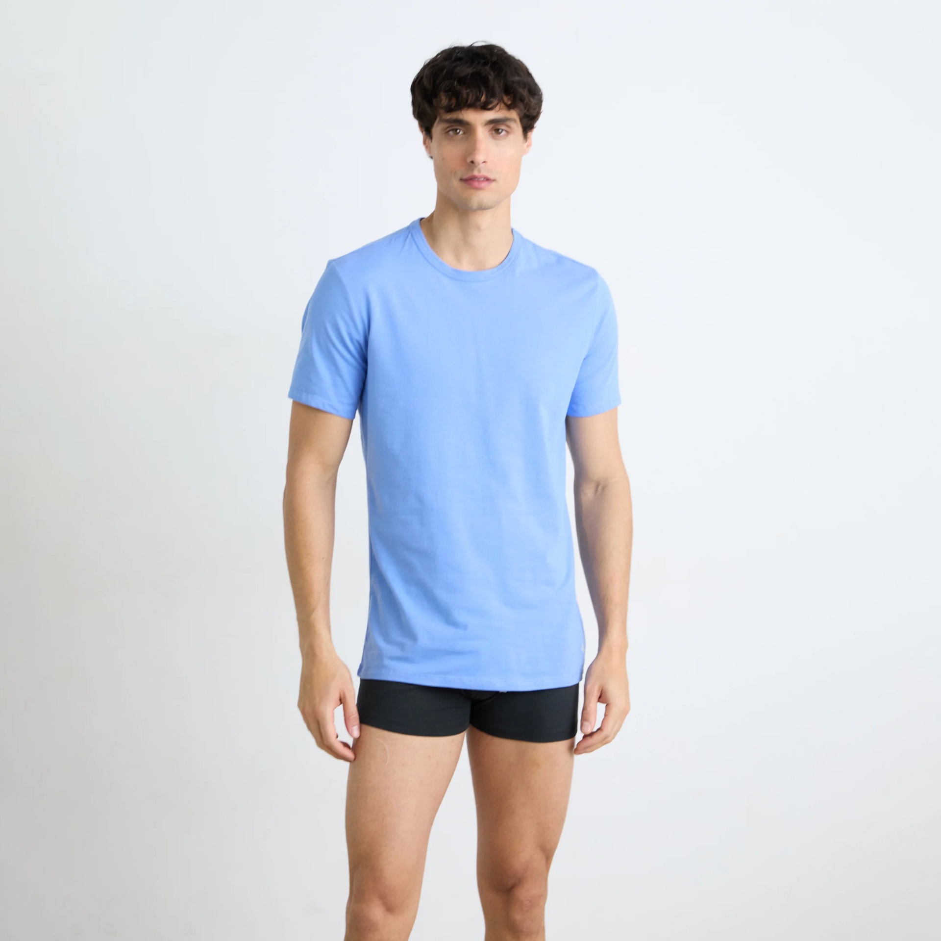 Homem com t-shirt azul clara de manga curta e cuecas pretas em fundo branco