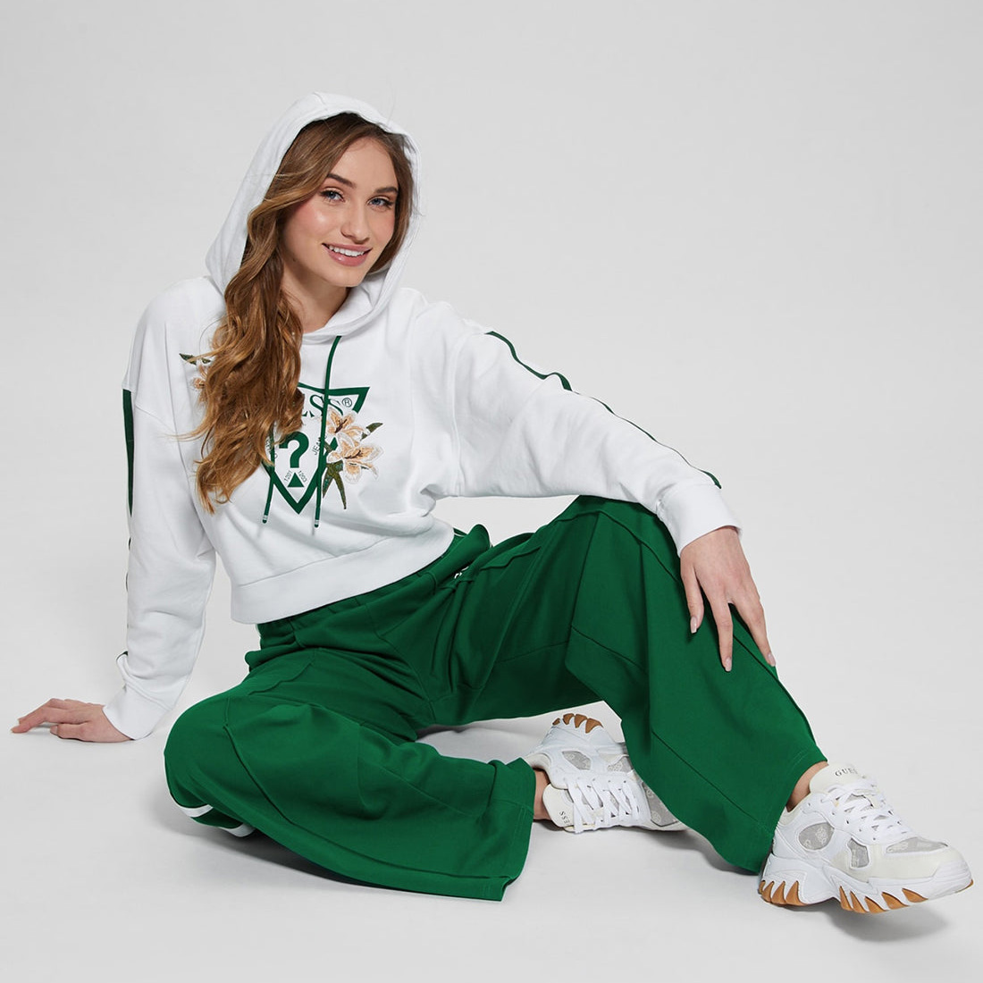 Mulher sentada com hoodie branco com logótipo triangular e flores, calças verdes largas e ténis brancos.