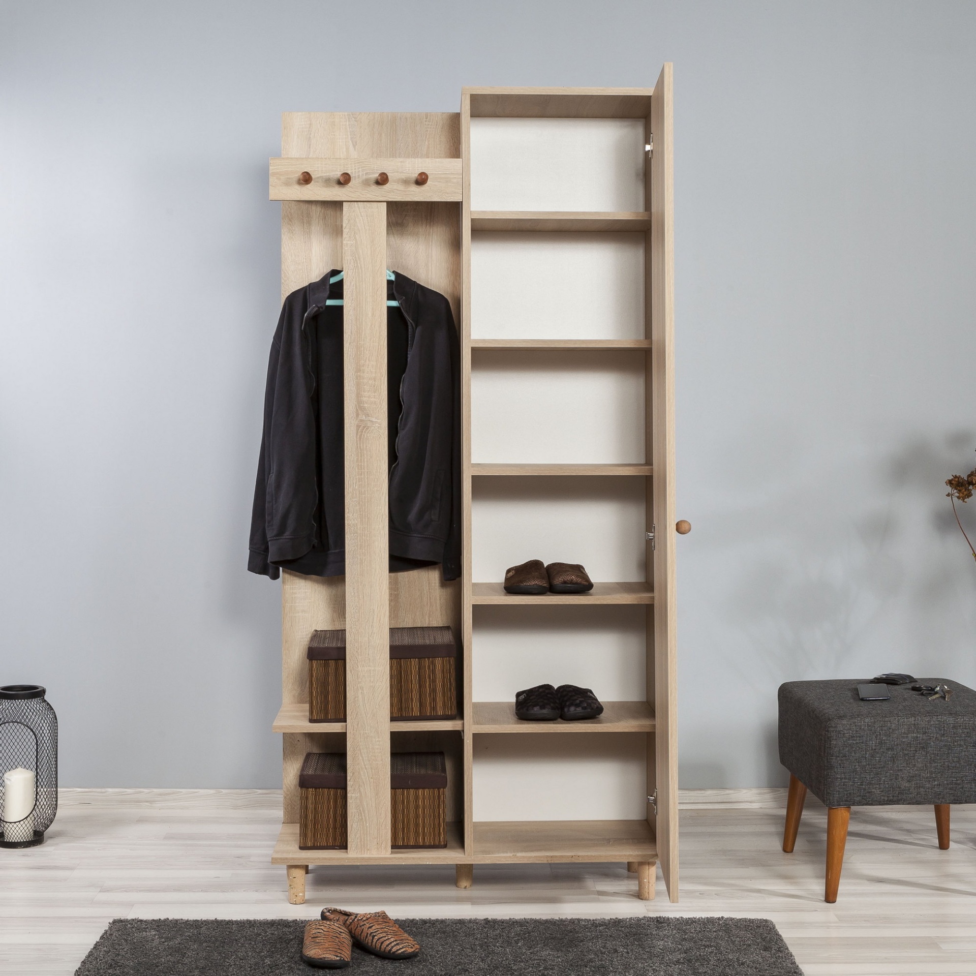 Armário de madeira clara com casaco e prateleiras com chinelos e caixas, ao lado tapete cinzento e banco cinzento.