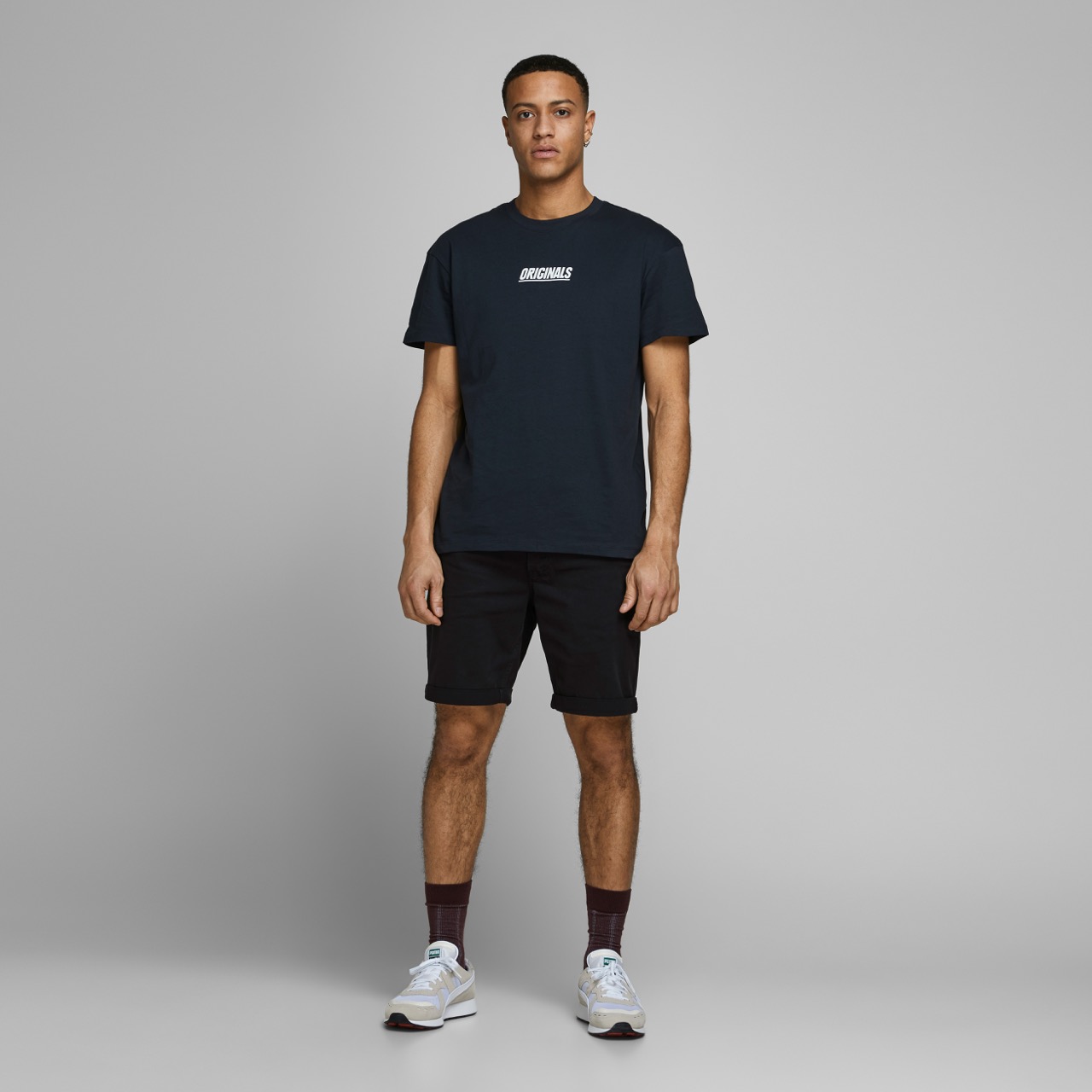 Homem com t-shirt preta com texto branco REGULAR, calções pretos e sapatilhas brancas em fundo cinzento.