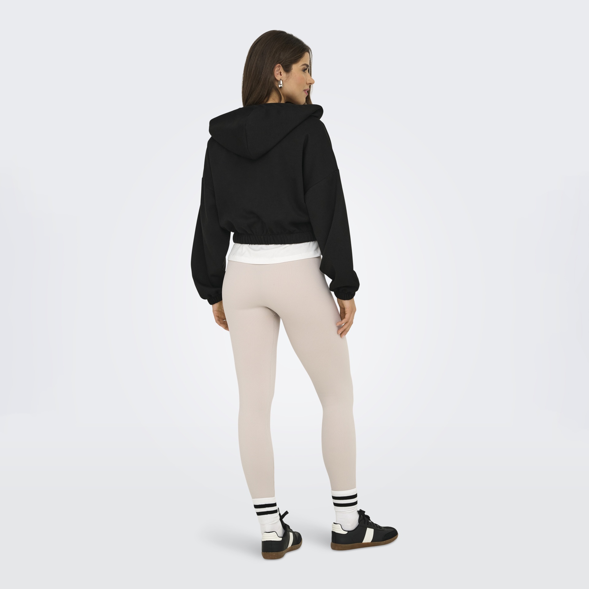 Mulher de costas com hoodie preto, leggings bege, meias brancas e sapatilhas pretas.