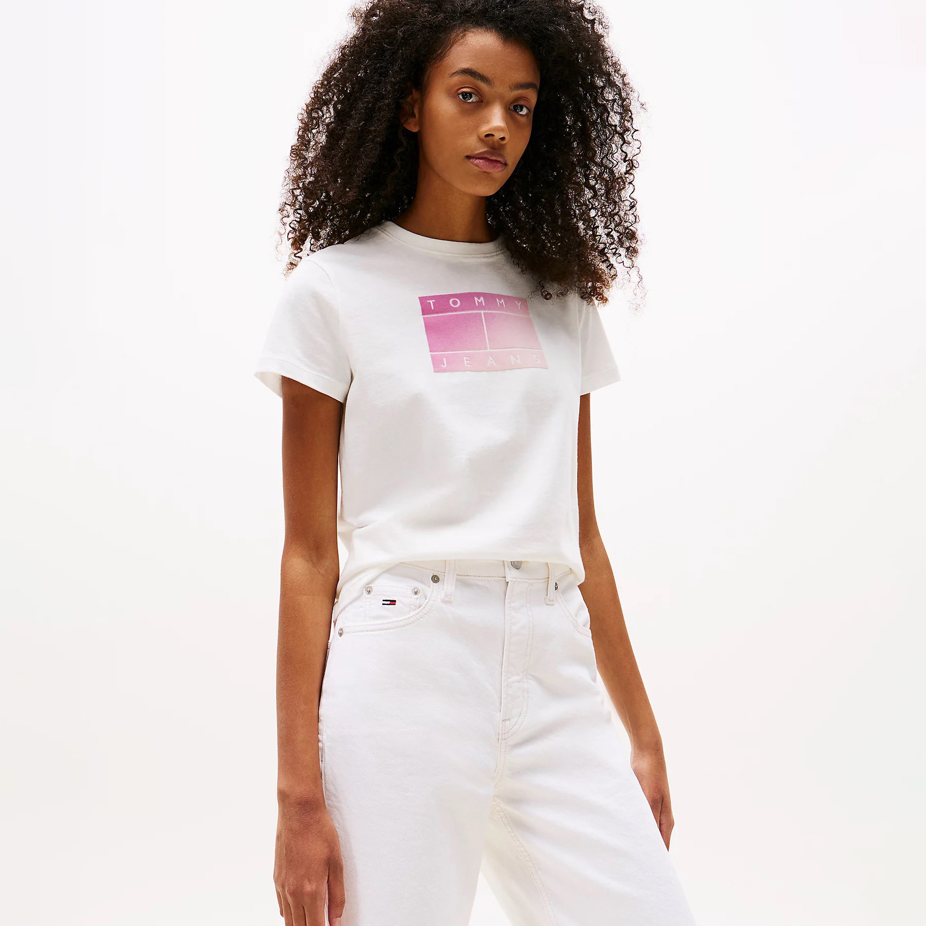 Modelo com camisola branca de manga curta com estampado rosa TOMMY JEANS e calças brancas de ganga com logotipo no bolso.