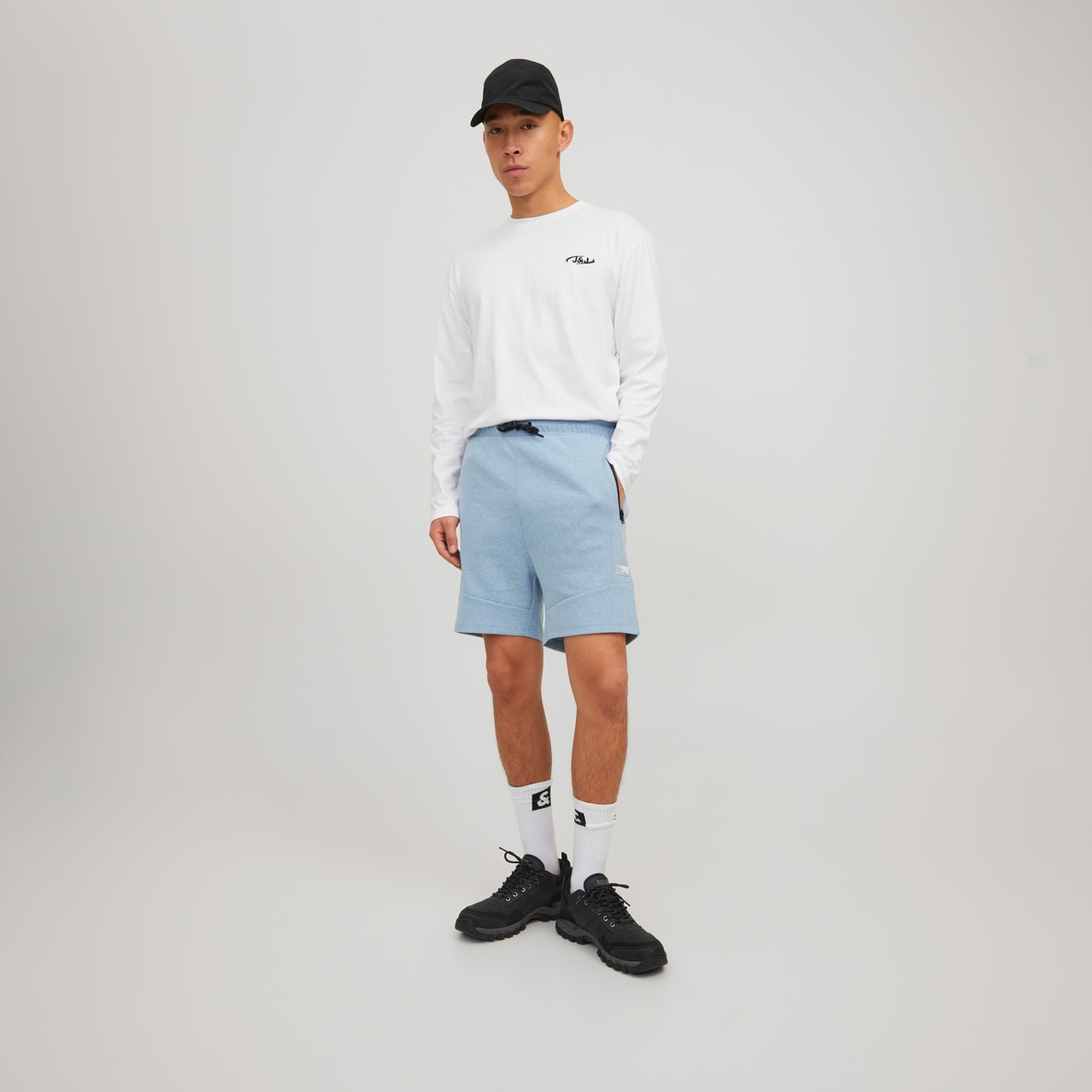 Homem com camisola branca, calções azuis, meias brancas, ténis pretos e boné preto