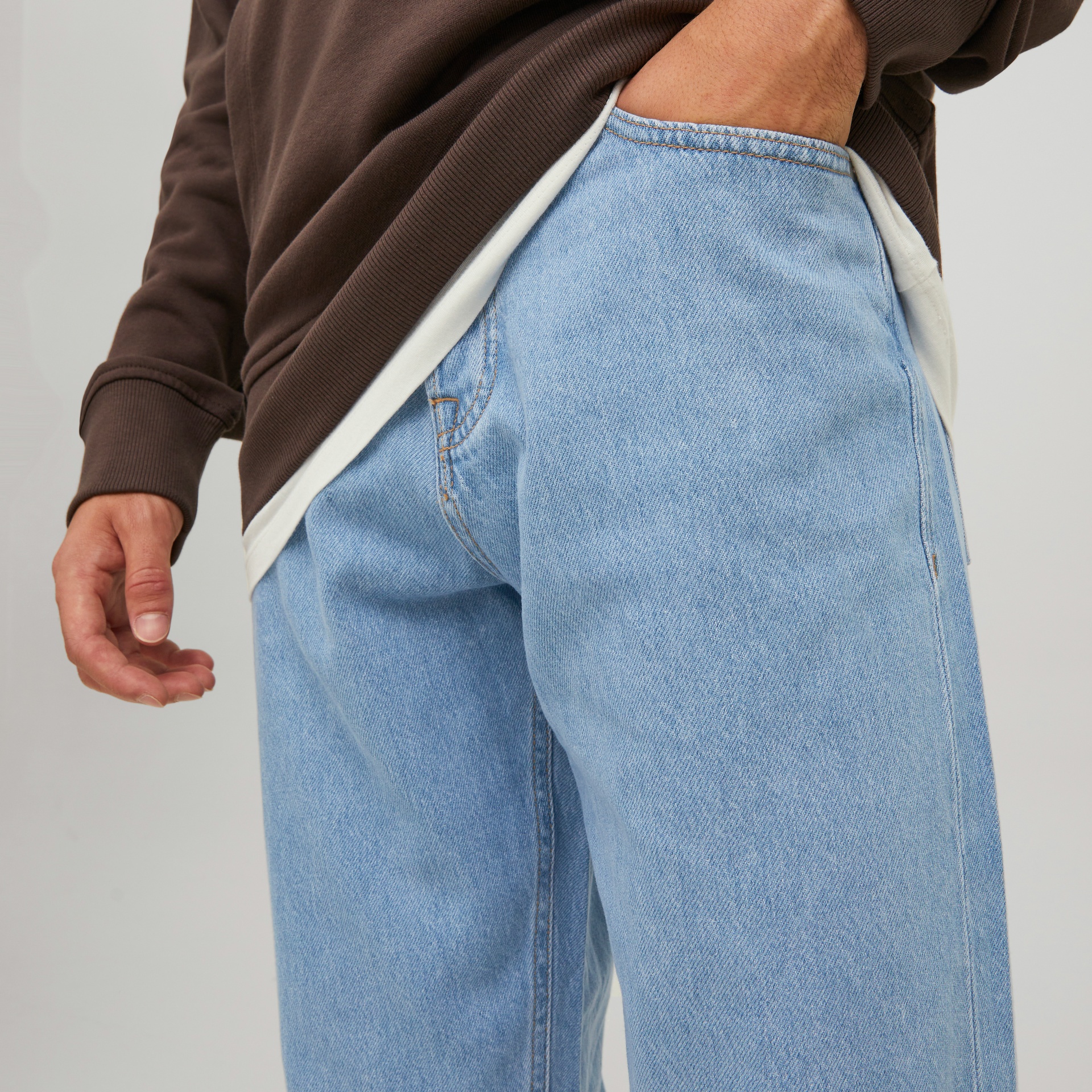 Calças de ganga azul claro com bolso lateral, parte inferior do suéter castanho e camisola branca visível