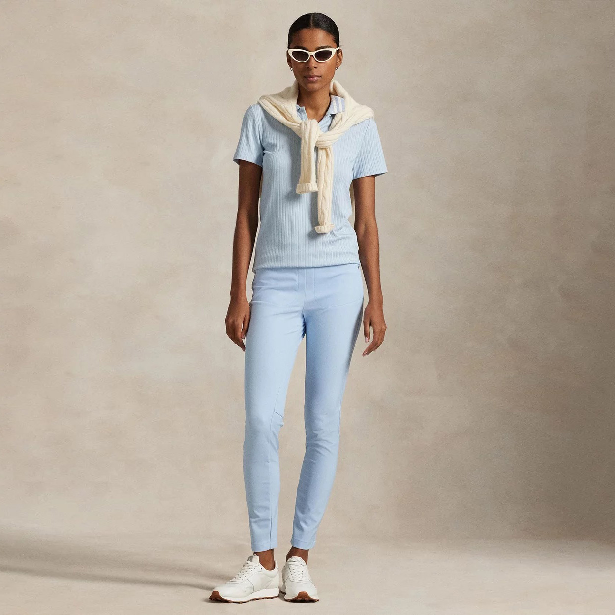 Modelo vestindo roupa casual azul claro e bege com óculos de sol brancos em fundo neutro