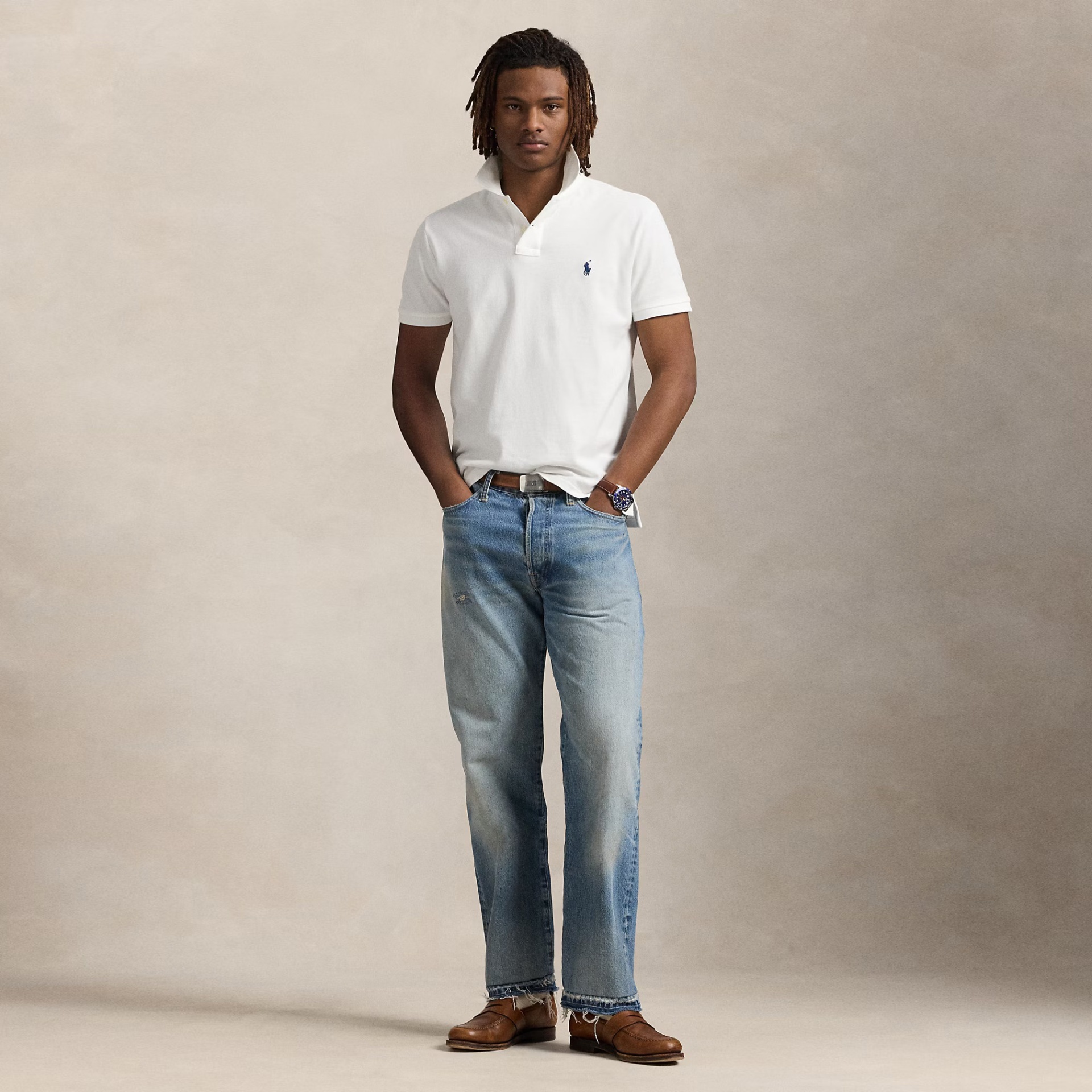 Homem de pé com pólo branco, jeans azuis claros e sapatos castanhos contra fundo neutro cinzento.