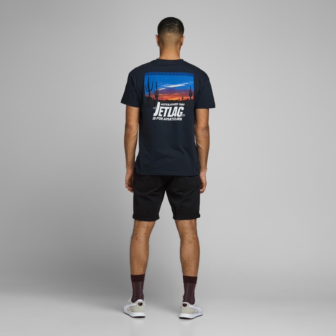 Homem de costas a usar t-shirt preta com imagem e texto nas costas, calções pretos, meias castanho escuro e sapatilhas brancas