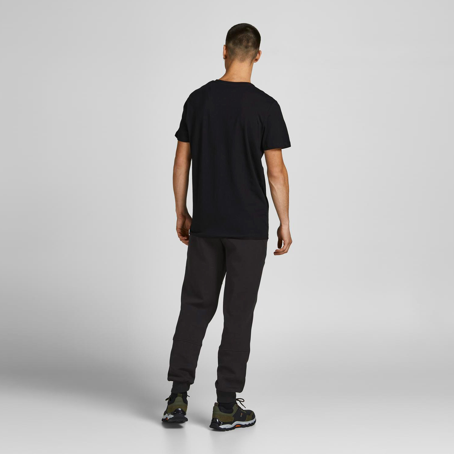 Homem com t-shirt preta, calças pretas e sapatilhas desportivas, visto de costas num fundo cinzento.