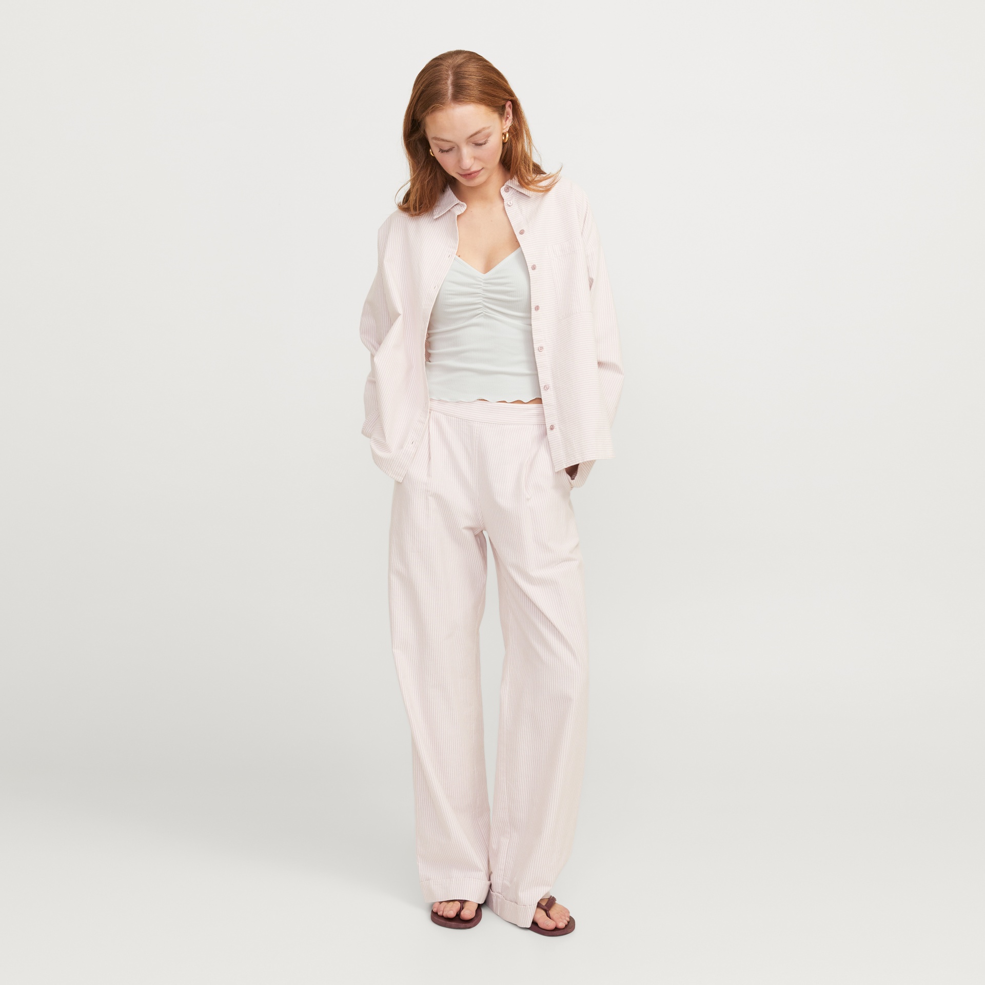 Mulher com conjunto riscado rosa claro, top branco e chinelos castanhos em fundo branco