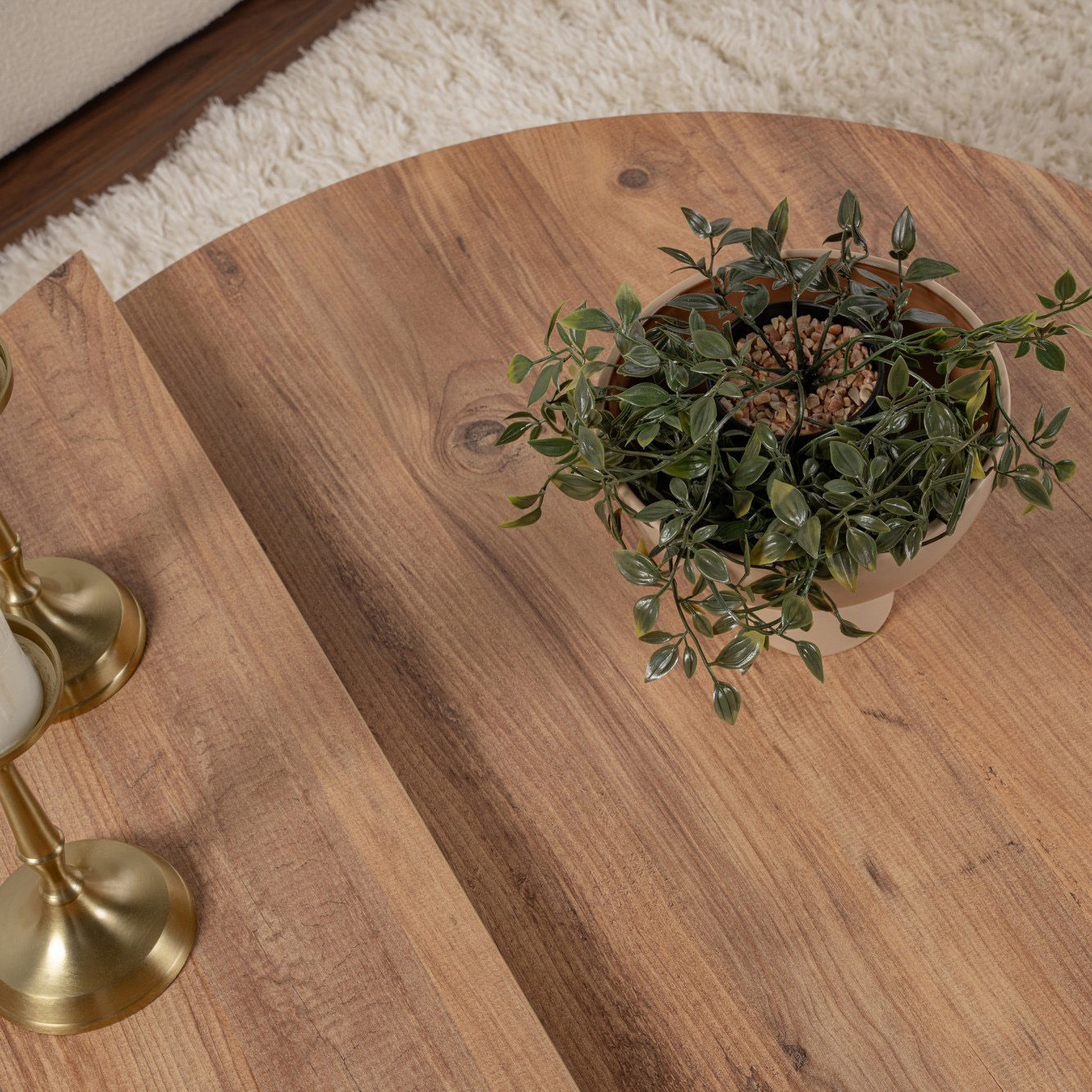 Mesa redonda de madeira clara com vaso de planta artificial verde e castiçais dourados num tapete creme