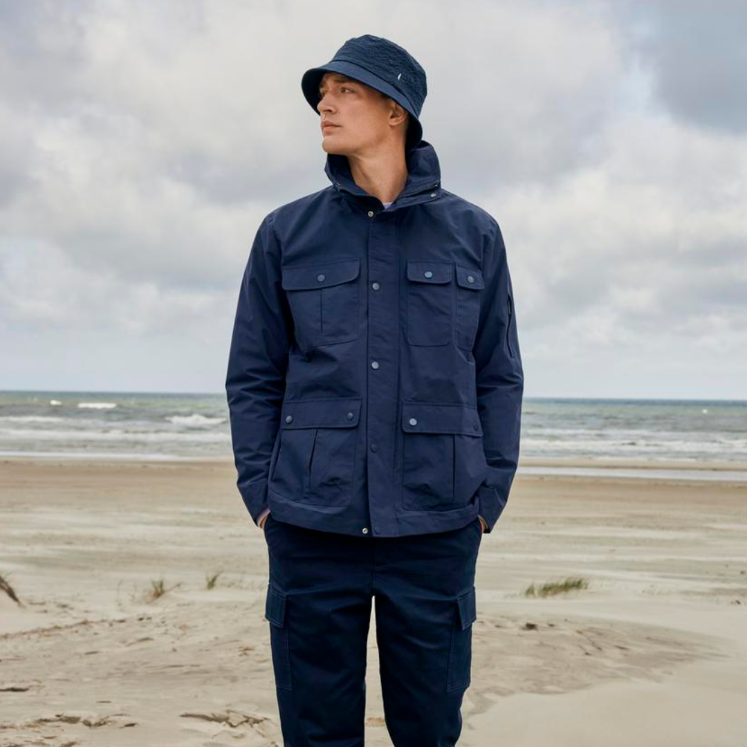 Homem com casaco e chapéu azul escuro na praia com mar e céu nublado