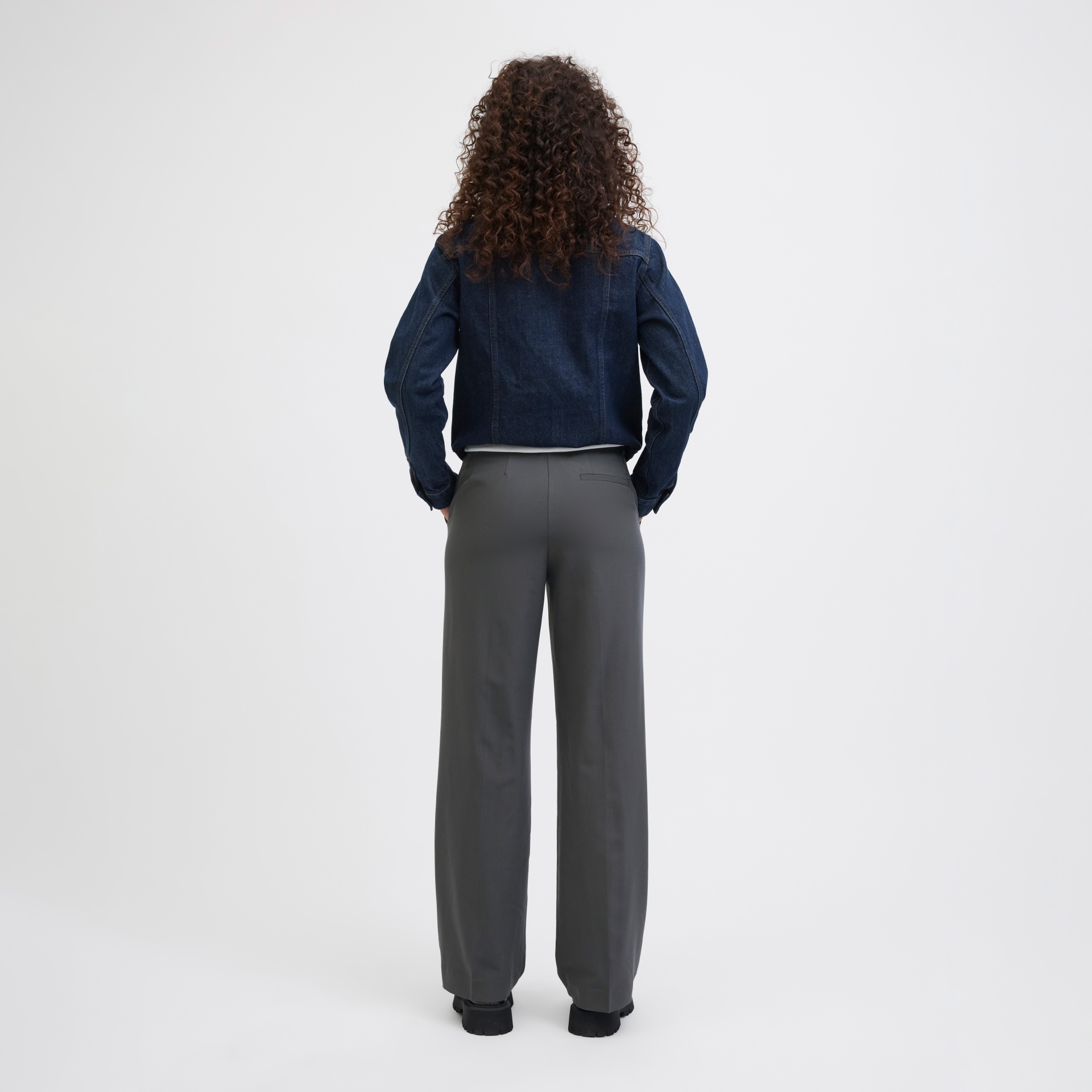 Pessoa de costas com casaco de ganga azul escuro, calças largas cinzentas e sapatos pretos em fundo branco.