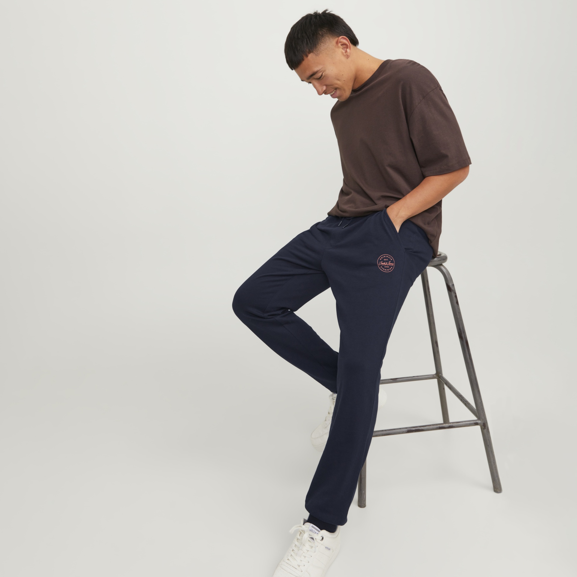 Homem sentado usando camisola castanha, calças de treino azuis com logótipo Jack & Jones e ténis brancos sentado num banco metálico.