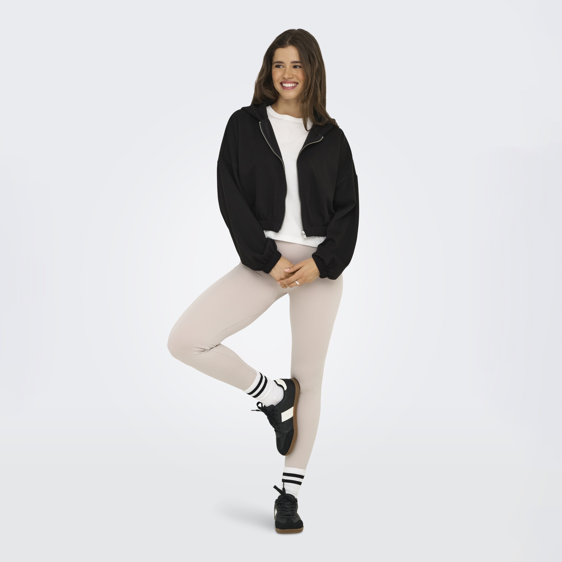 Mulher a usar casaco preto com fecho e capuz, t-shirt branca, leggings bege e sapatilhas pretas