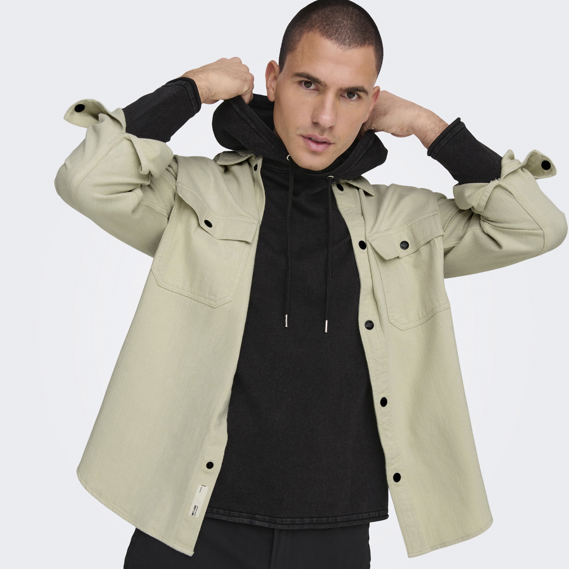 Homem veste camisola com capuz preta e casaco bege claro com bolsos ao peito