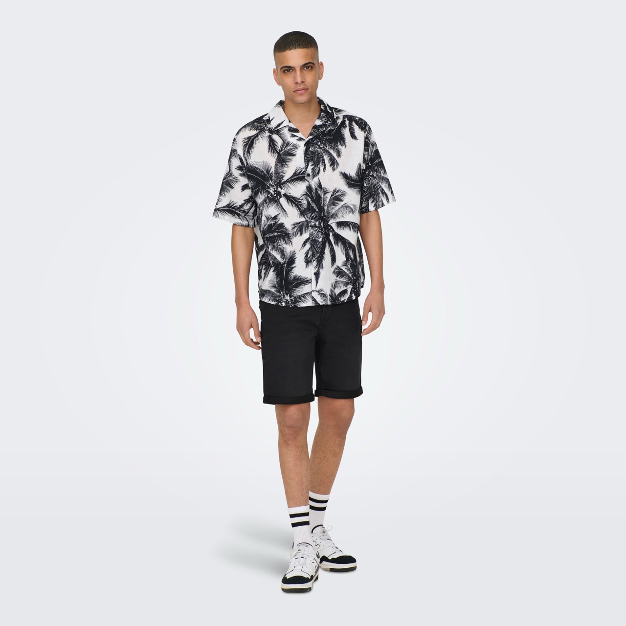 Homem vestido com camisola preta e branca com padrão de folhas e calções pretos, meias brancas às riscas e ténis brancos.