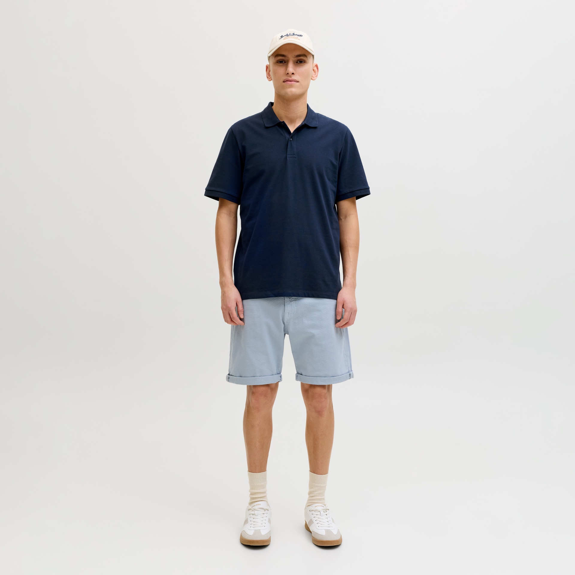 Homem com polo azul escuro, calções azuis claros, boné bege e sapatilhas brancas