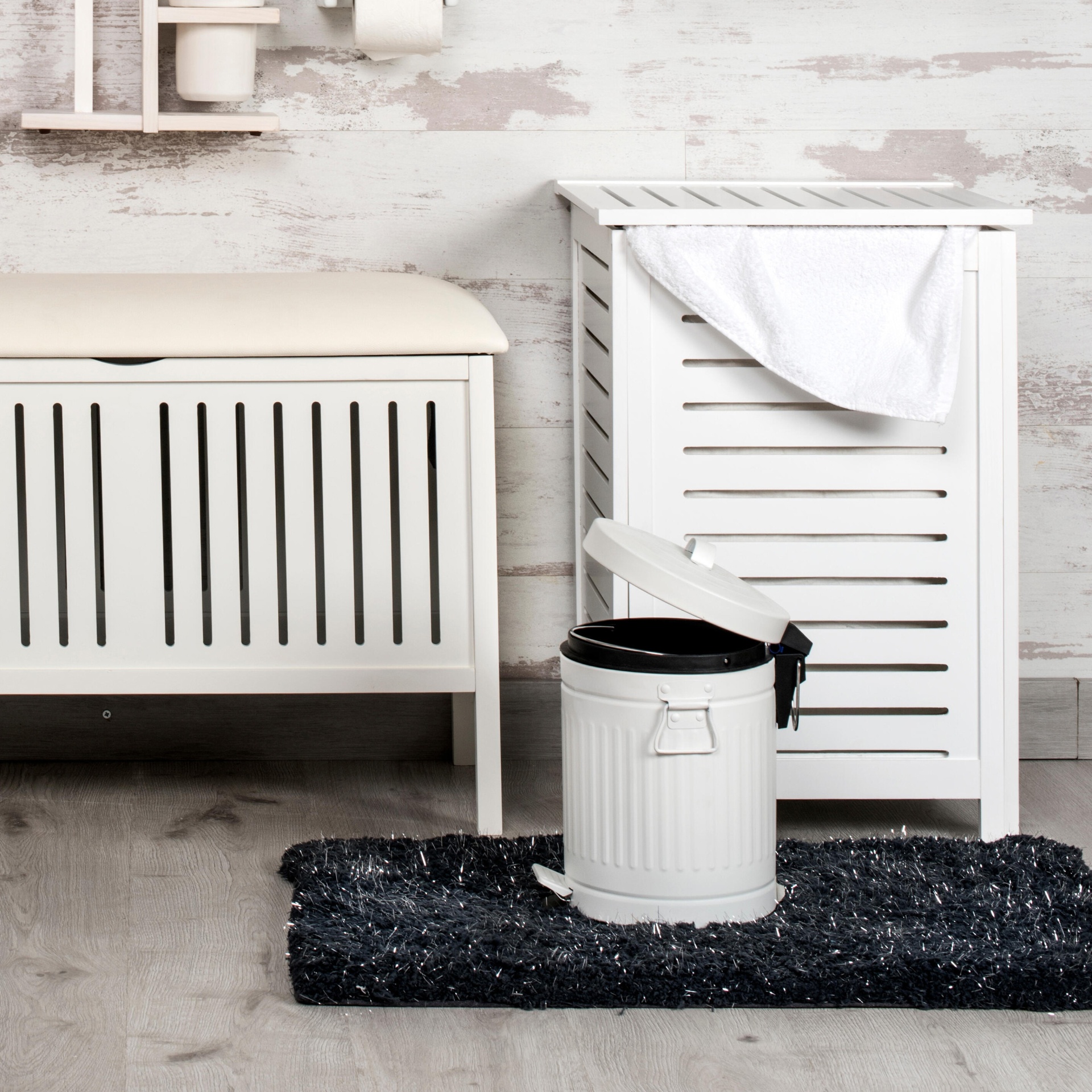 Móveis de WC brancos com banco almofadado, armário de ripas, toalha branca e balde de lixo metálico branco sobre tapete preto.