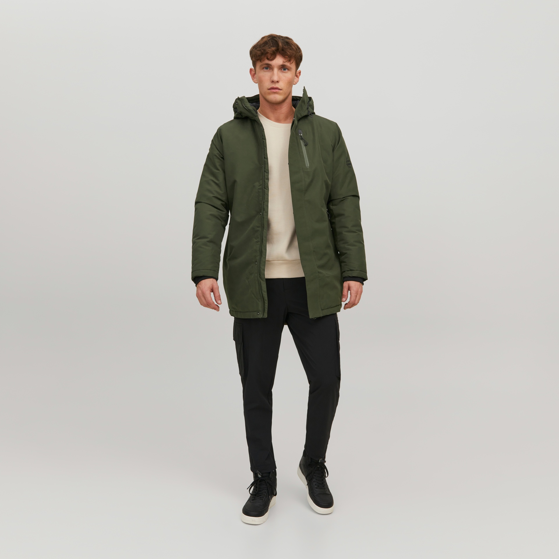 Homem com casaco verde, camisola bege, calças pretas e sapatilhas pretas