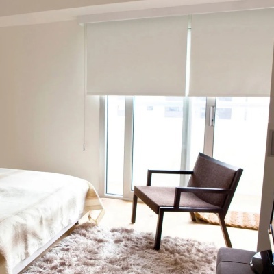 Quarto com cama branca, cadeira castanha, tapete peludo e persianas brancas