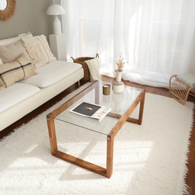 Mesa de centro de vidro e madeira numa sala iluminada junto a sofá creme e cortinas brancas