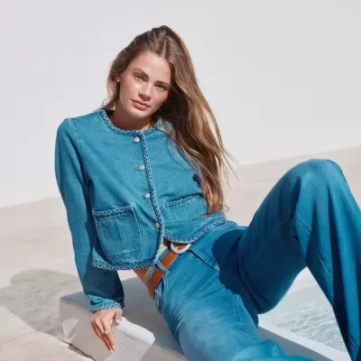 Casaco de ganga azul com detalhes trançados e botões prateados usado por mulher sentada.