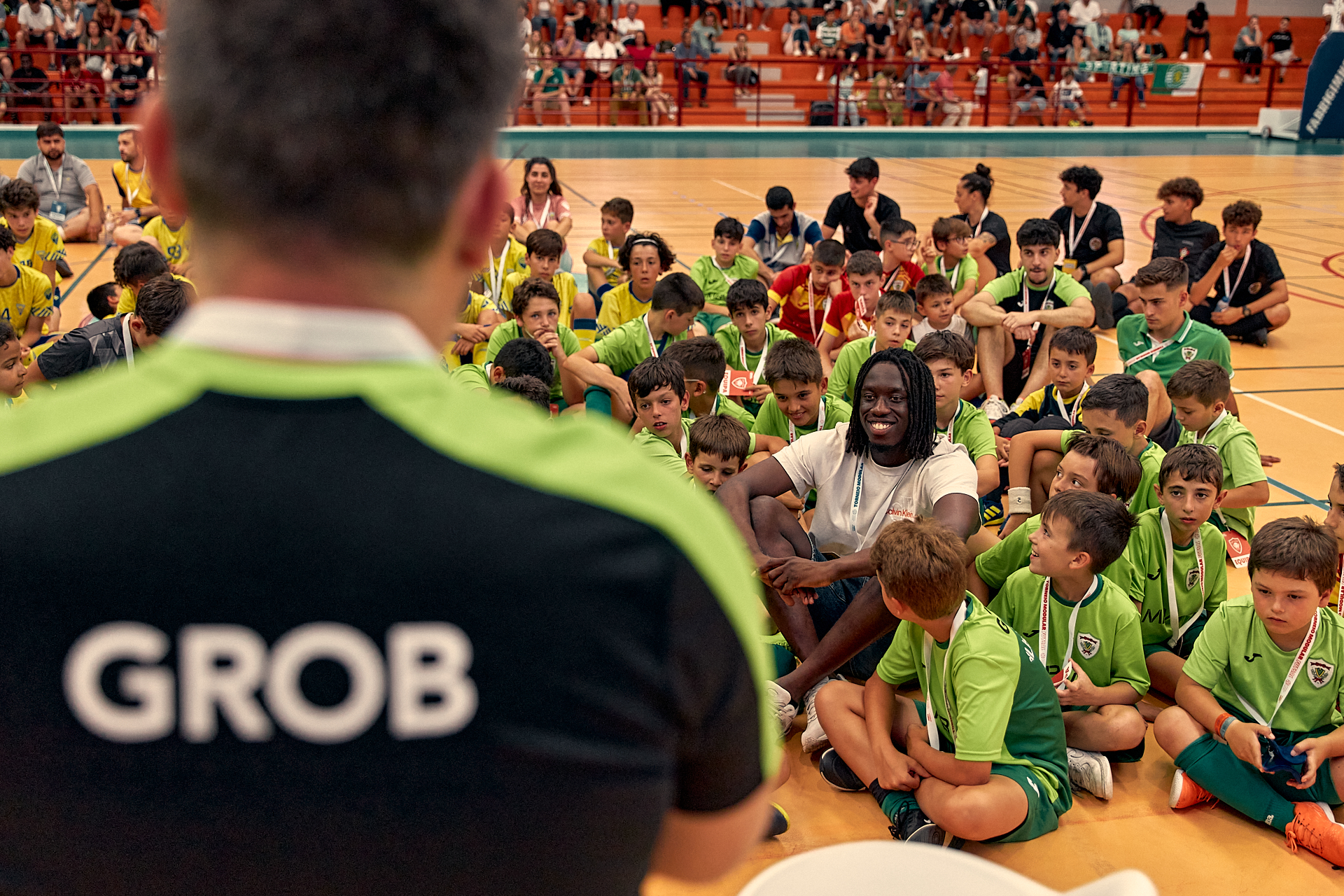 Camisola preta e verde com GROB nas costas em ginásio com crianças em uniformes verdes e amarelos sentadas no chão