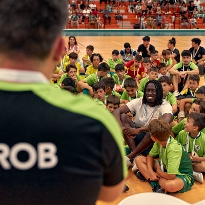 Camisola preta e verde com GROB nas costas em ginásio com crianças em uniformes verdes e amarelos sentadas no chão