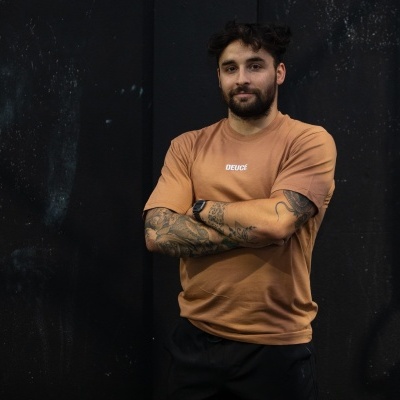 Homem com t-shirt castanha DEUCE, tatuagens nos braços e fundo negro texturizado