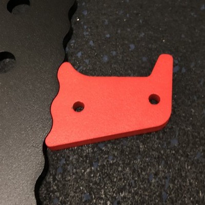 Peça metálica vermelha com dois orifícios sobre superfície preta texturizada ao lado de peça metálica preta com borda ondulada e furos