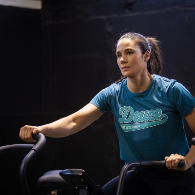 Mulher a usar bicicleta estática com t-shirt azul turquesa com texto 'Deuce NEW YORK 1987'