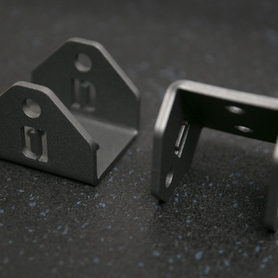 dois suportes metálicos cinza escuro com furos para fixação sobre superfície escura pontilhada