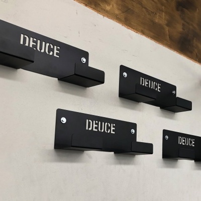 Suportes de parede pretos com texto 'DEUCE' para pendurar, fixados em parede branca, com cordas grossas ao lado.