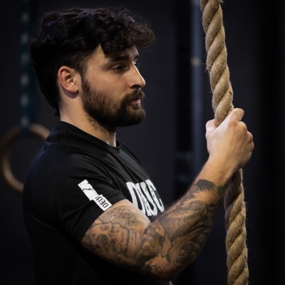 Homem com tatuagens segurando corda em ginásio com argolas de madeira ao fundo
