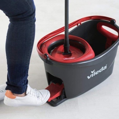 Balde de limpeza Vileda cinza e vermelho com pedal e esfregona ao lado de perna com calças de ganga e sapatos brancos.