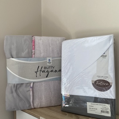 Conjunto de cama embalado com etiquetas visíveis e cobertor cinzento com detalhe floral sobre superfície de madeira