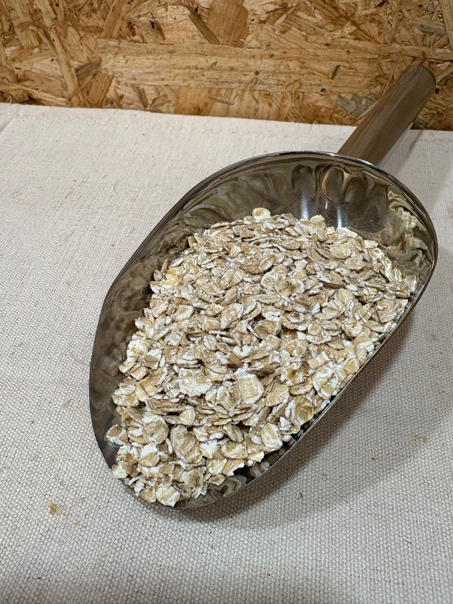 Flocos Aveia Finos Pá metálica com flocos de aveia crus e cabo de madeira sobre tecido branco e fundo de madeira OSB