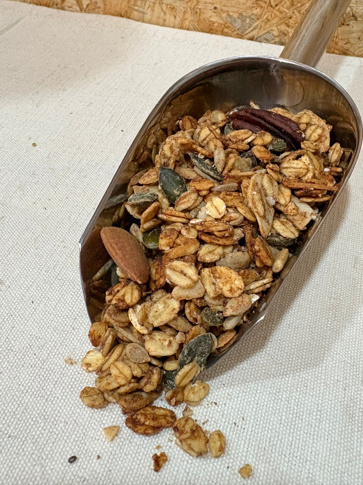 Granola Manteiga de Amendoim e Canela Colher metálica com granola mista contendo aveia, sementes e nozes sobre tecido bege claro
